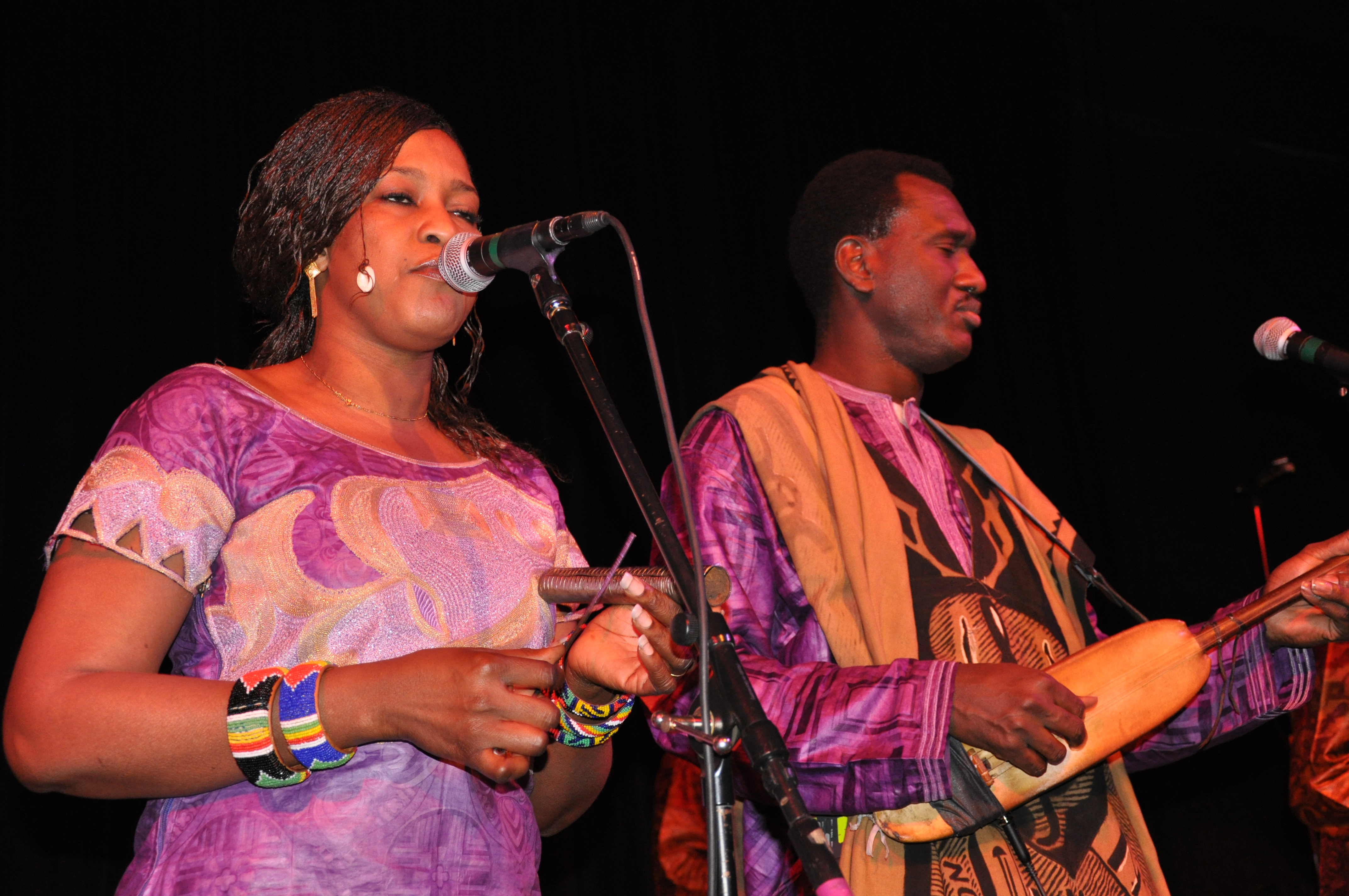 A woman singing into a microphone and a man playing a traditional stringed instrument on stage, both dressed in colorful african attire.