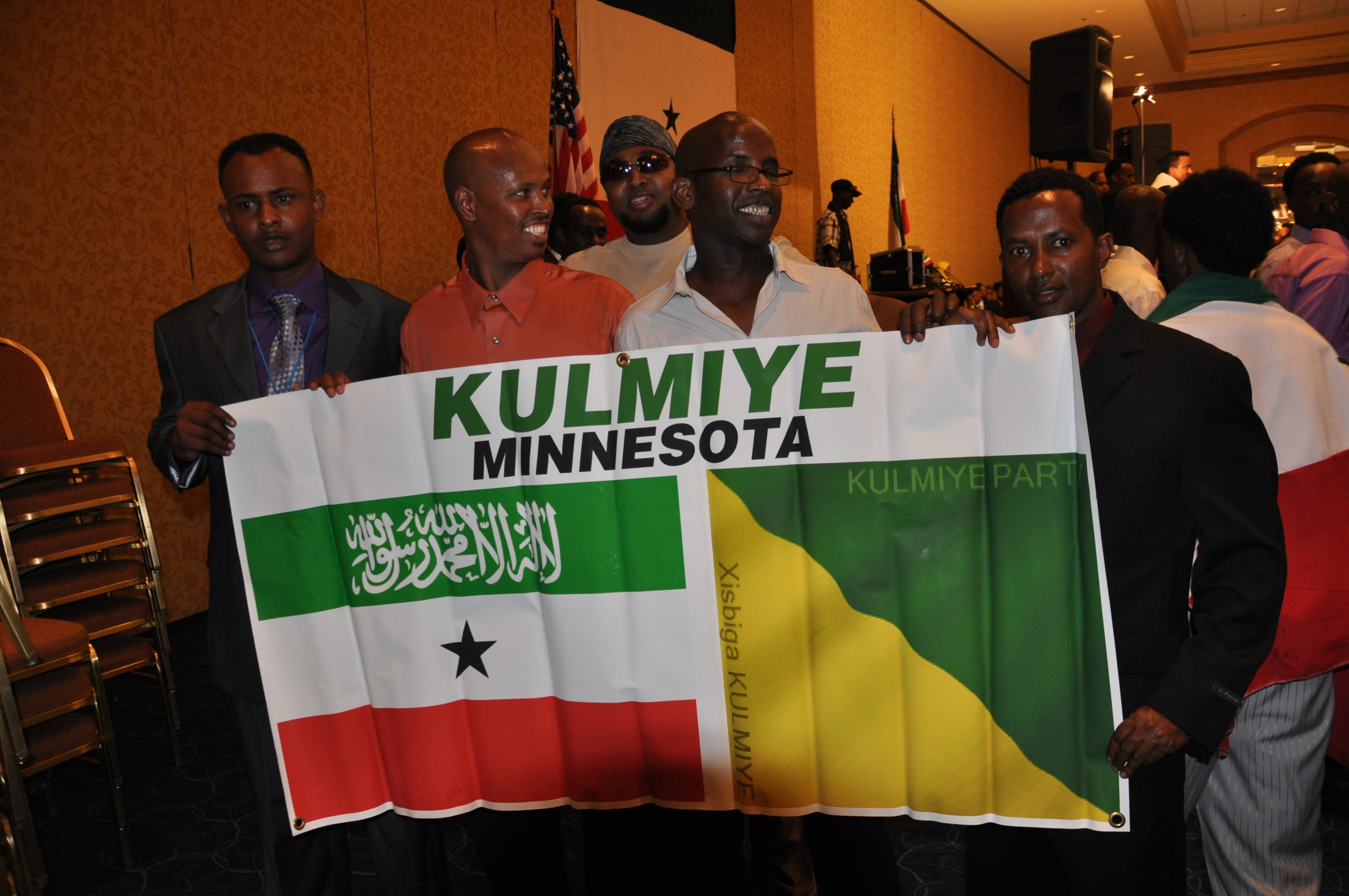 Five men standing, holding a large kulmiye party flag with the somaliland national flag colors and text, in a room with american flags in the background.