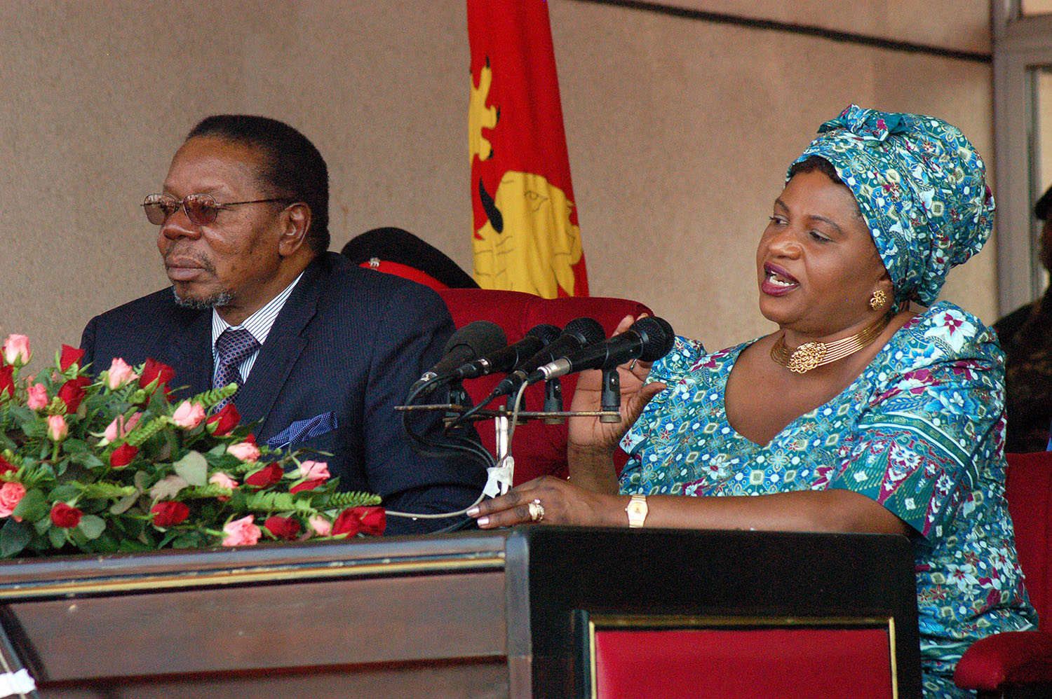 A man and a woman seated at a podium adorned with flowers; the woman is speaking into a microphone, both are dressed in formal attire.