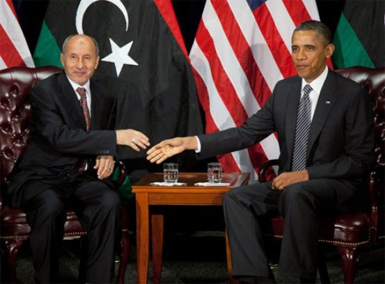 Two men shaking hands at a table with the flags of libya and the usa in the background.