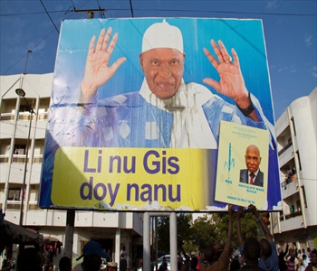 People holding up a large billboard featuring a smiling man in religious attire with raised hands, and a smaller sign with another man's portrait, in a busy street setting.