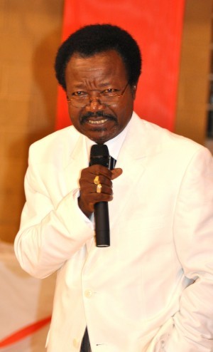 A man with a mustache, wearing a white suit and holding a microphone, speaks at an indoor event.