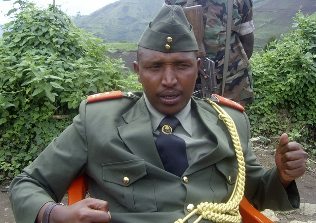 A man in a green military uniform with decorative cords sits outdoors, looking directly at the camera with a serious expression.