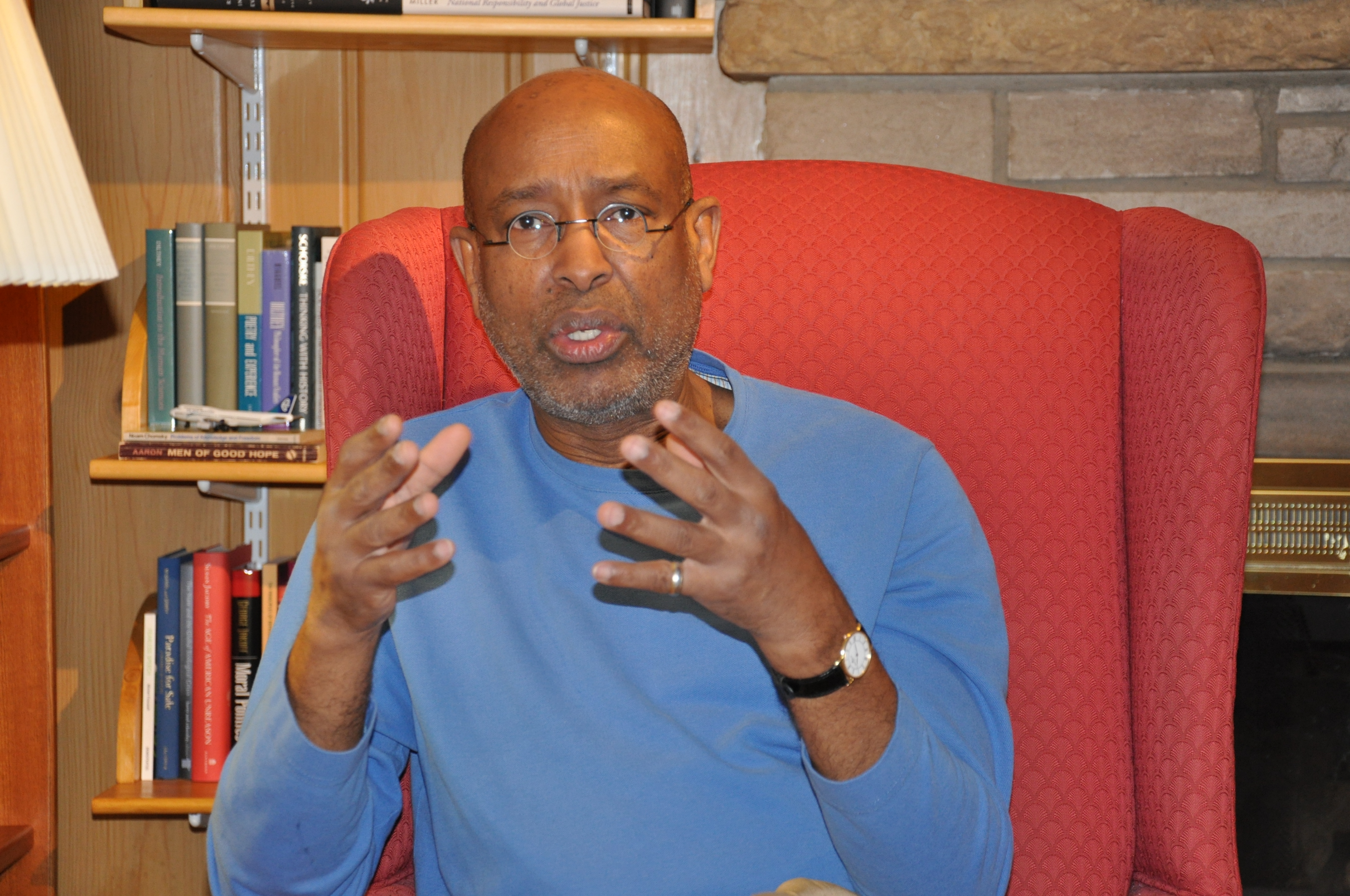A man with glasses wearing a blue sweater gestures while speaking, seated in a red armchair in a room with books.