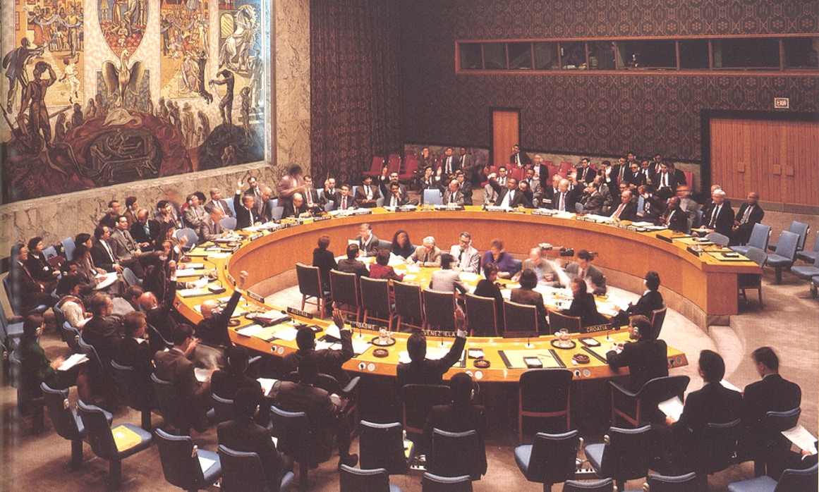A conference room with officials seated around a large oval table, engaging in discussions, with a colorful tapestry in the background.