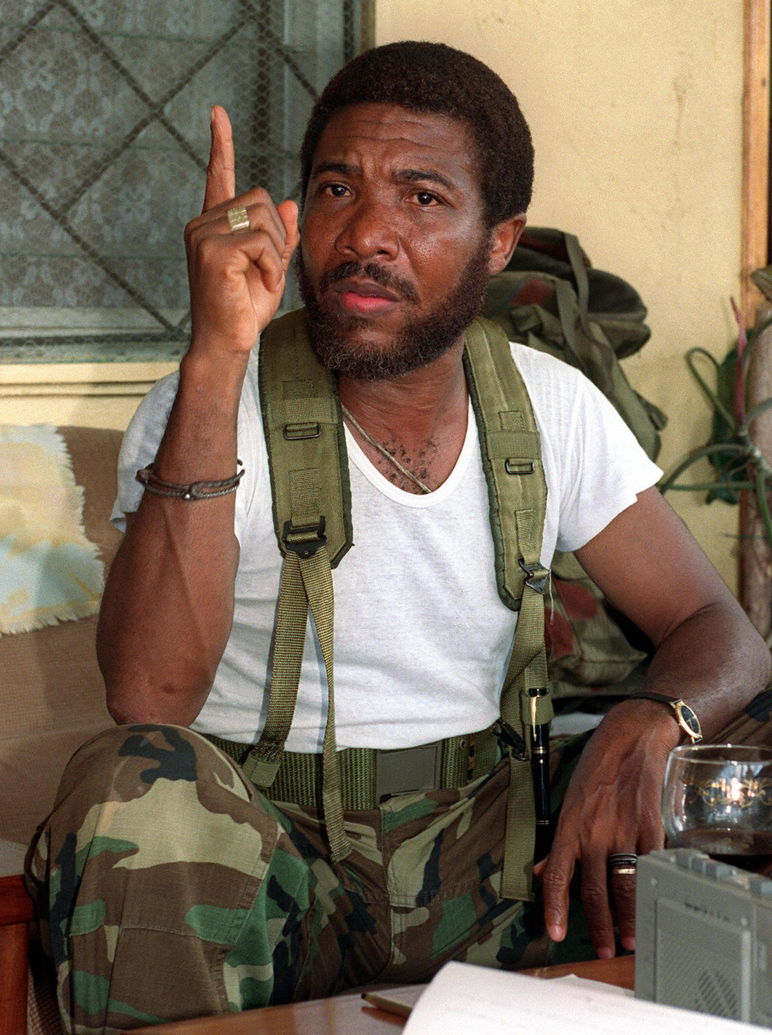 A man in military camouflage vest gestures with one finger raised, sitting in front of a patterned curtain, with a microphone visible in the foreground.