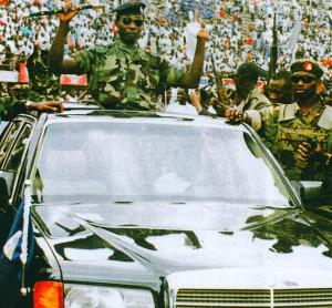 A military officer in uniform salutes from an open car in a parade, surrounded by other uniformed officers, with a large crowd in the background.
