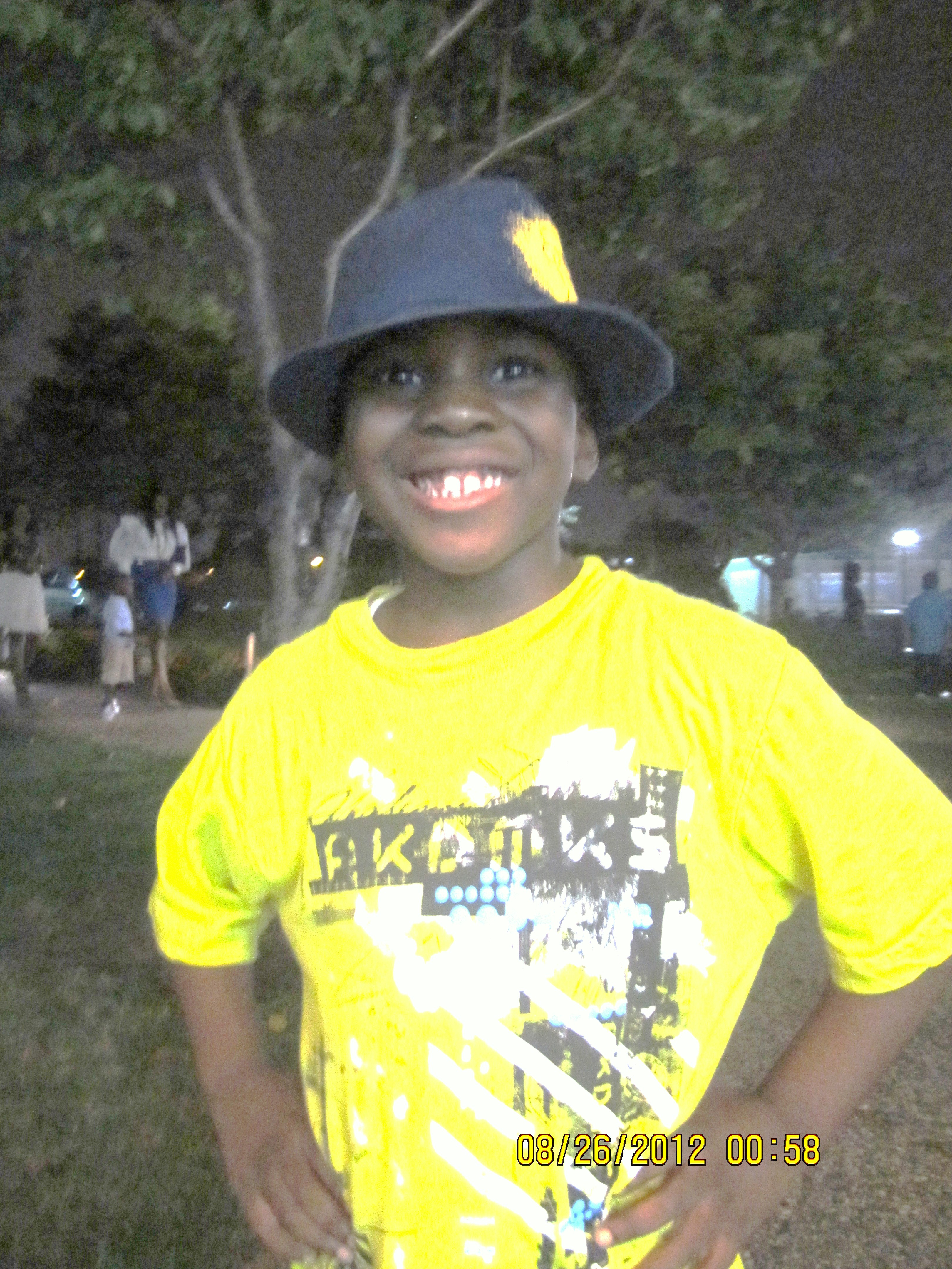 A young boy smiling at night, wearing a yellow graphic t-shirt and a blue hat, with trees and people in the background.