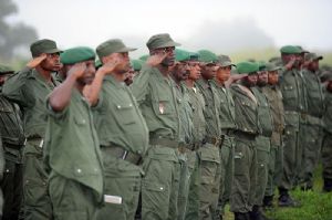 A line of soldiers in green uniforms and berets saluting solemnly during a misty outdoor ceremony.