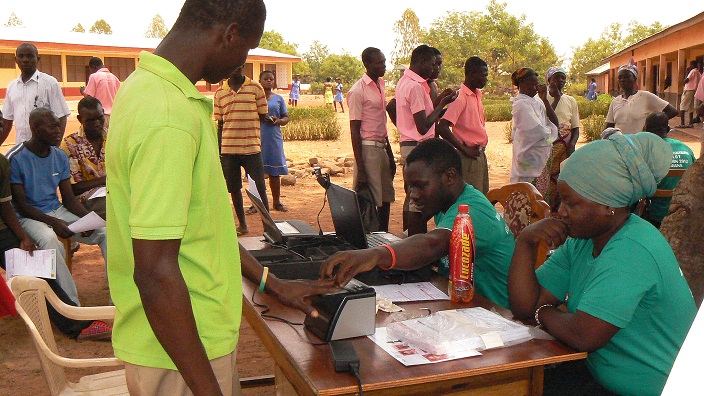 People at an outdoor registration event with laptops and documents on tables, interacting under a sunny sky.