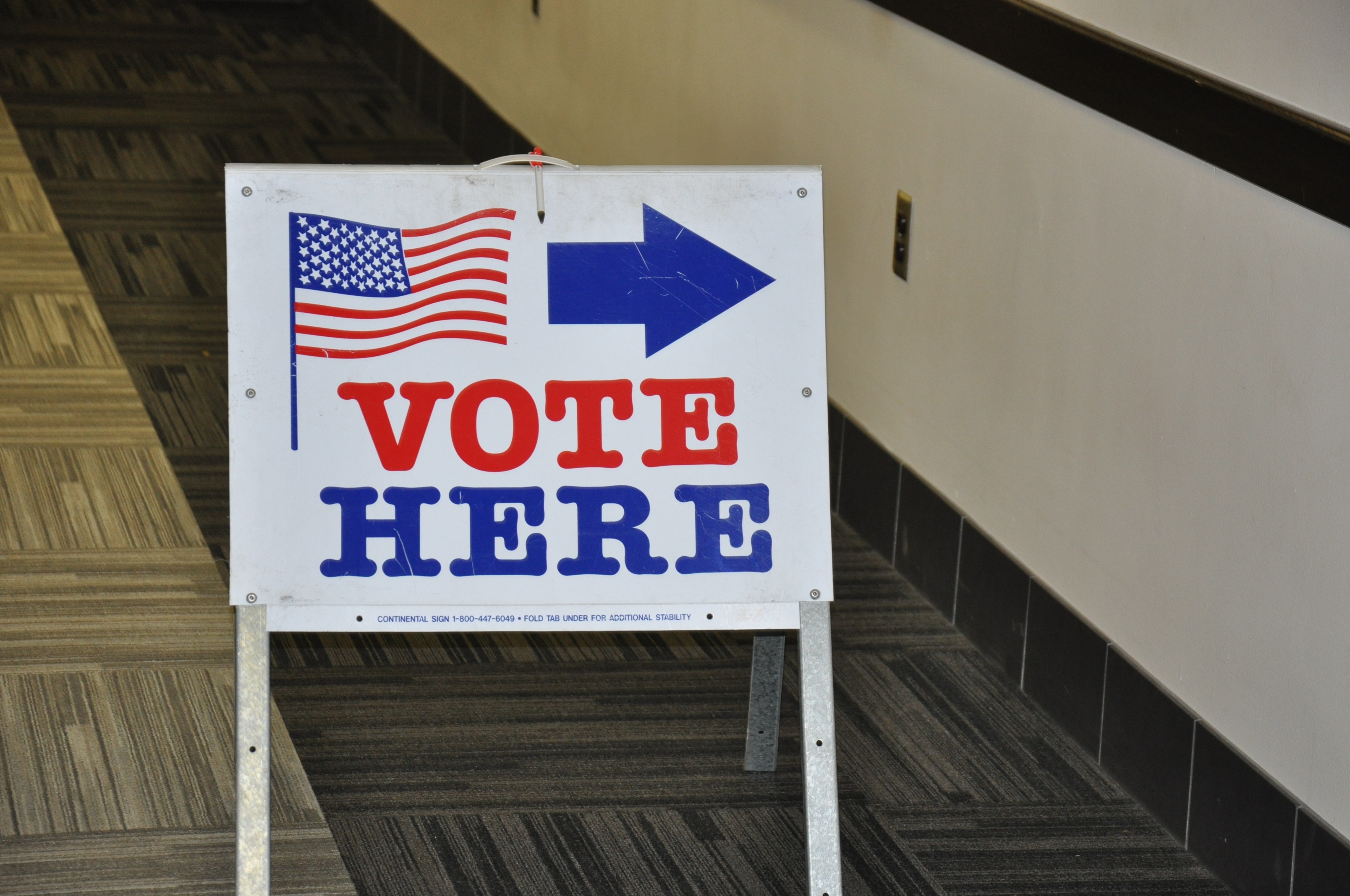A "vote here" sign with an american flag and a blue arrow pointing to the right, placed in a hallway with carpeted flooring.