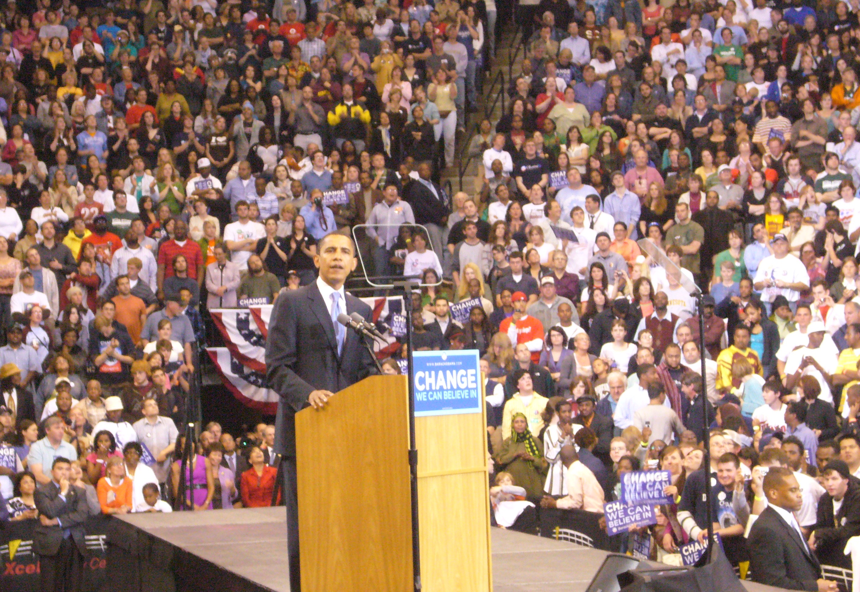 A man speaking at a podium with "change we need" banners in front of a large, diverse audience at an indoor arena.