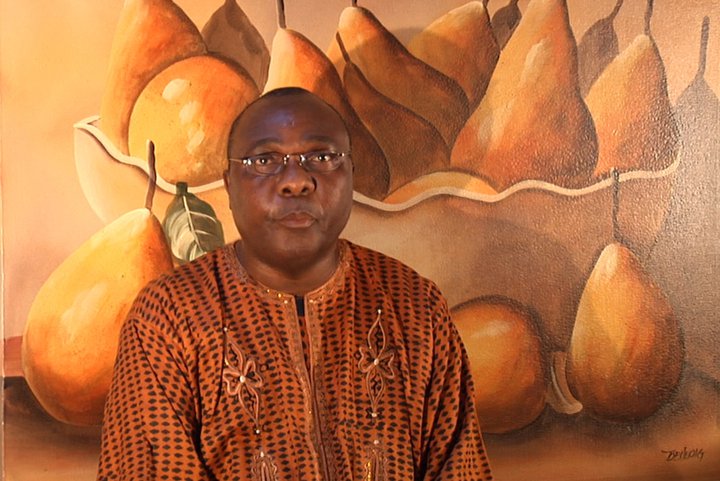 A man in traditional african attire poses in front of a painting featuring large, textured pears.