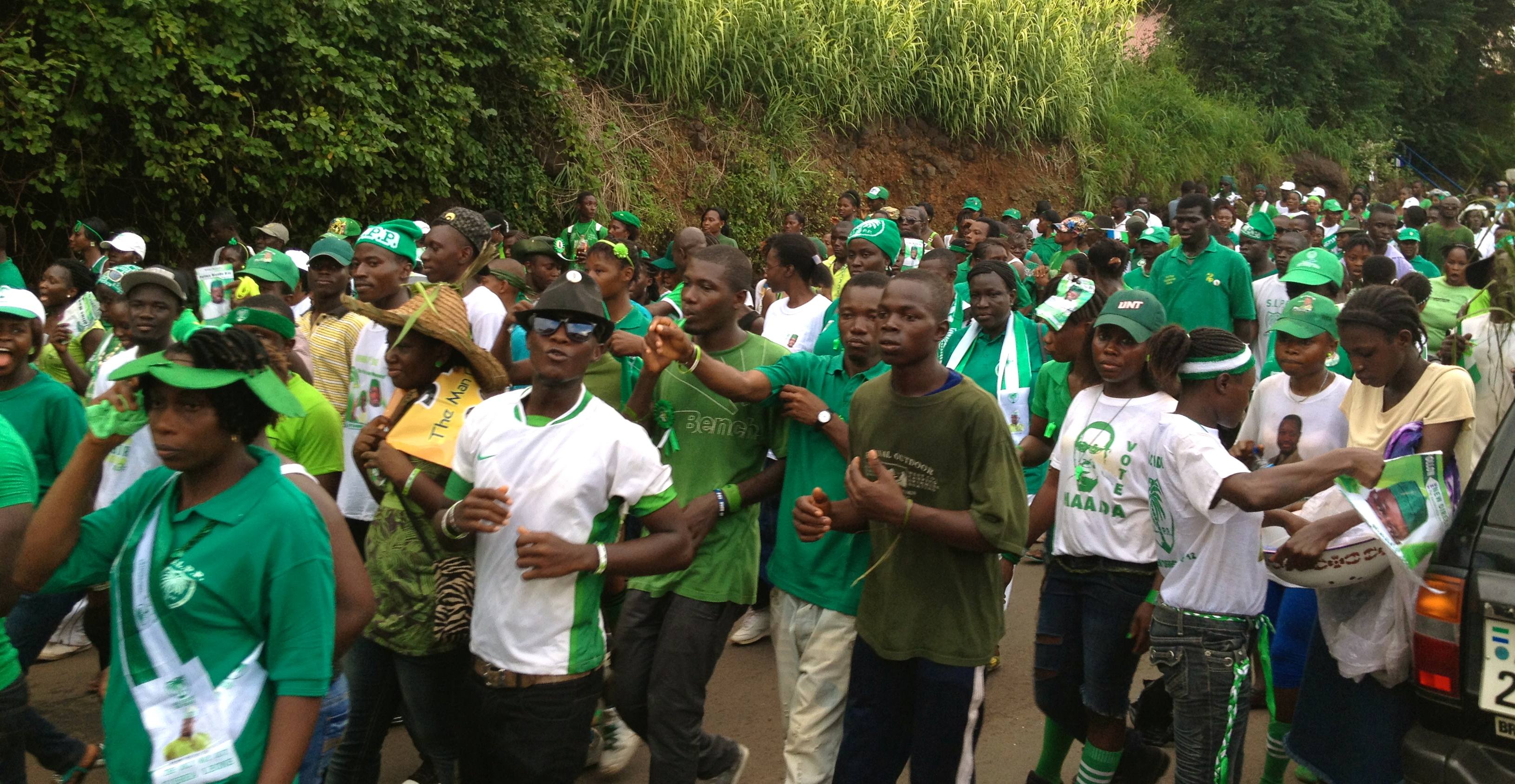 A large group of people wearing green outfits and caps, walking along a road during a rally or parade, holding banners and flags.