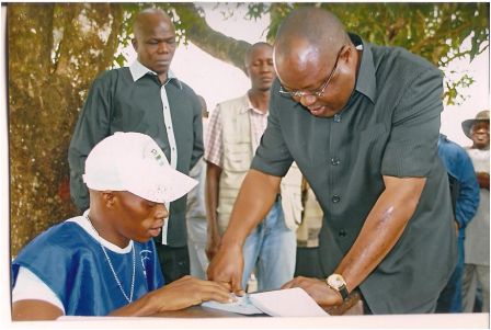 Two men standing under a tree, one observing the other as he signs a document, with several onlookers in the background.