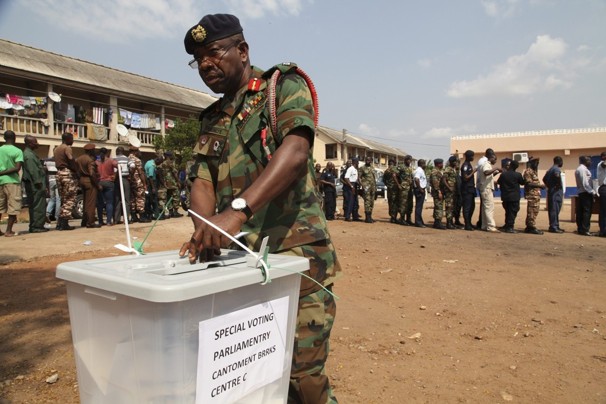 A military officer in uniform casting a vote into a transparent ballot box at a polling station, with observers and personnel in the background.