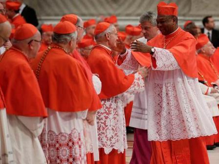 Cardinals in red and white vestments greeting each other inside a large hall.