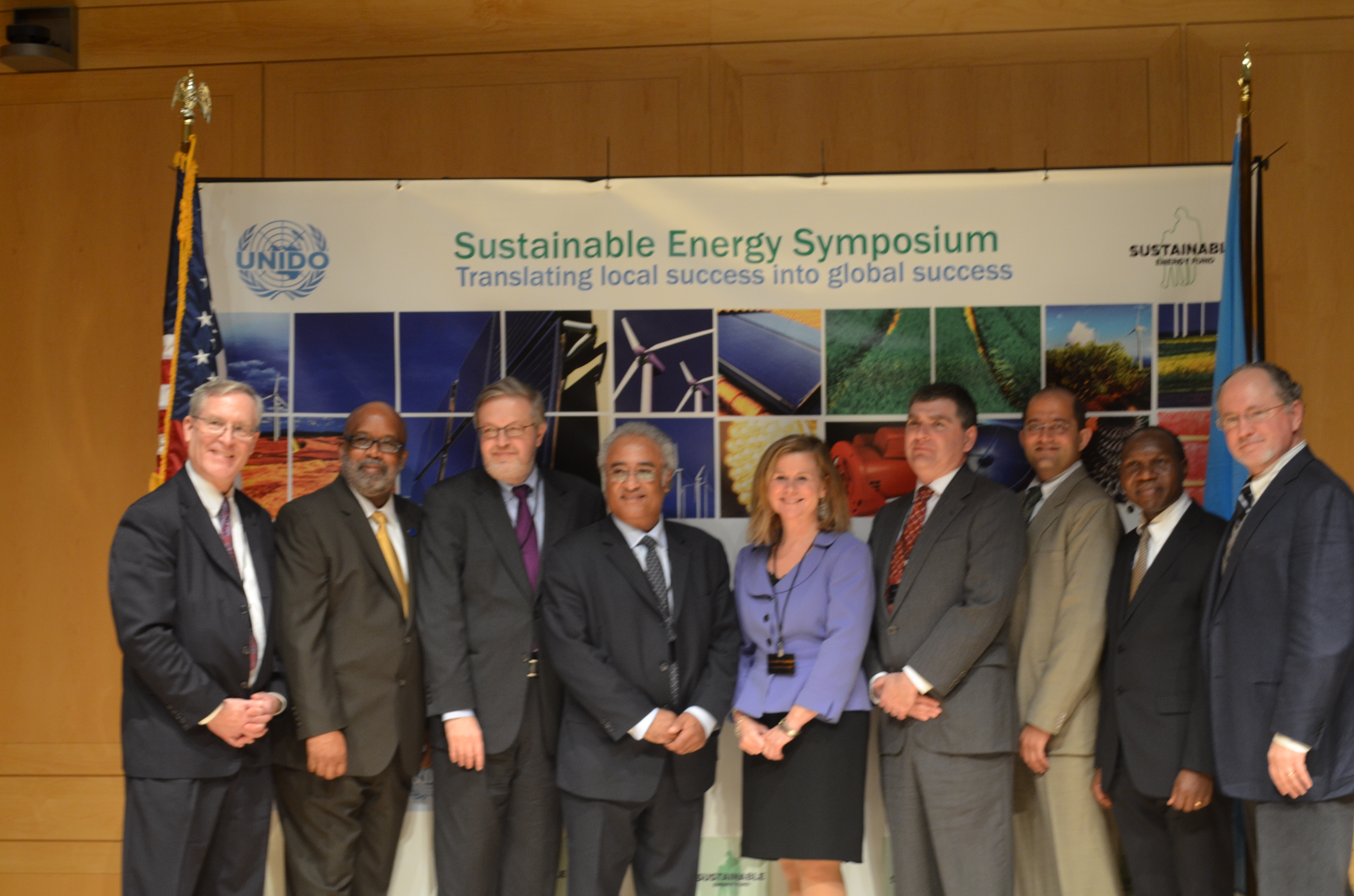 Group of nine people posing in front of a banner at the sustainable energy symposium, featuring images of wind turbines and solar panels.