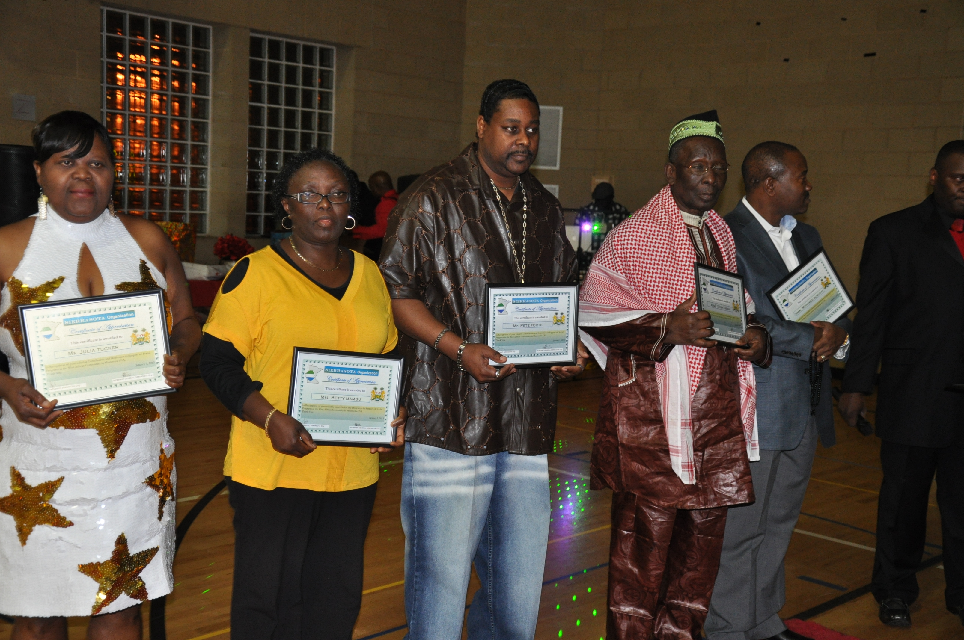 Five adults standing in a gym, each holding a certificate; three men and two women dressed in formal and traditional attire.