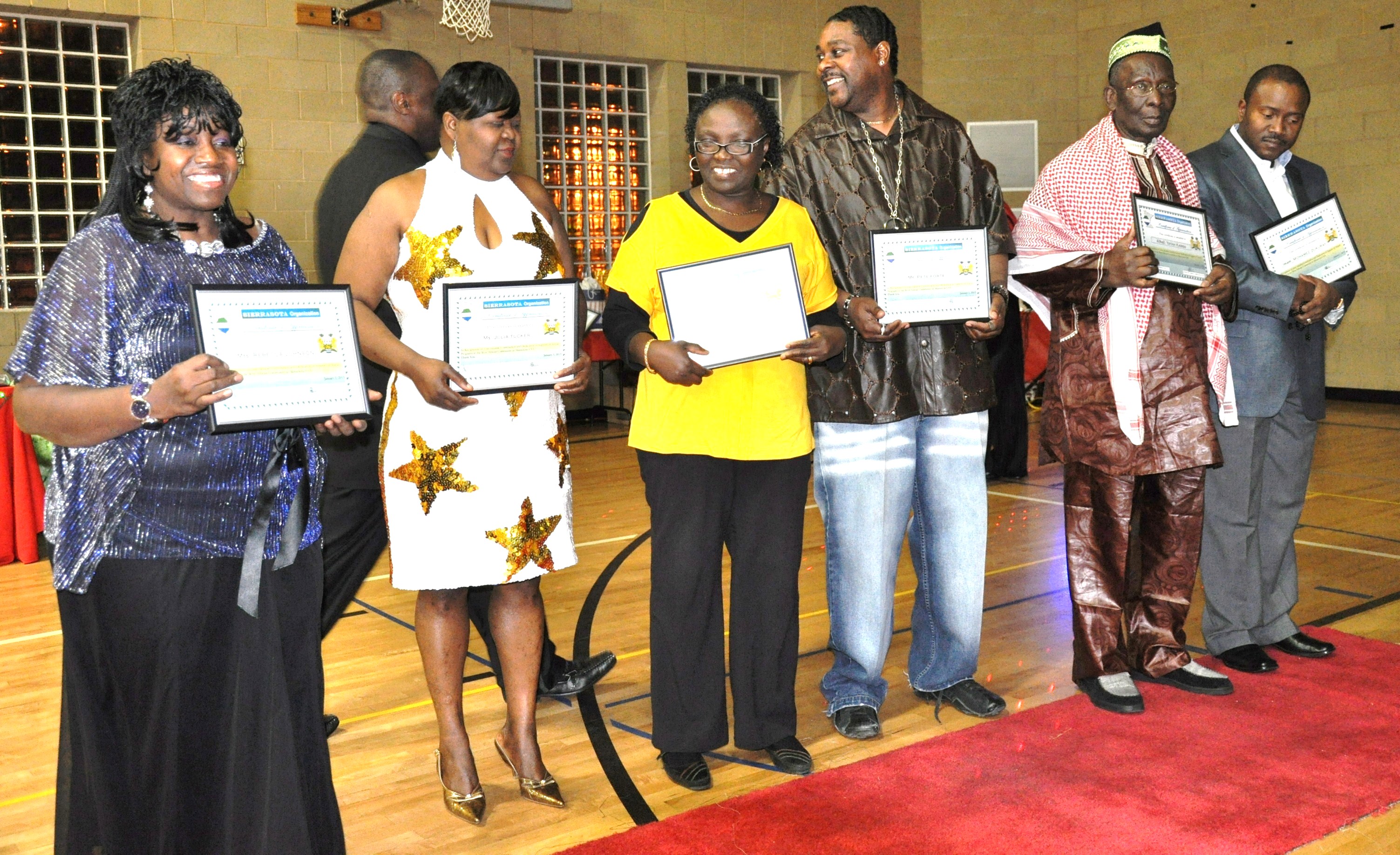 Group of six adults smiling and holding certificates in a gymnasium, dressed in formal and traditional attire.