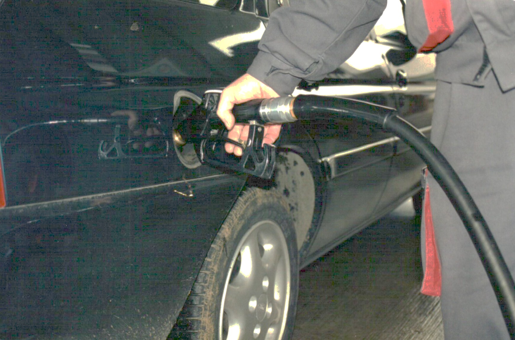 A person refueling a dark car at a gas station, focusing on the fuel nozzle inserted into the car's tank.