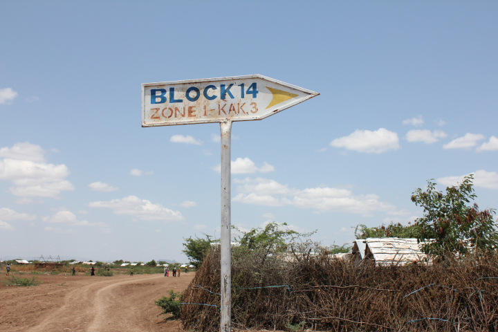 Directional sign labeled "block 14 zone 1 - kak 3" with a dusty path and thatched huts under a clear blue sky.