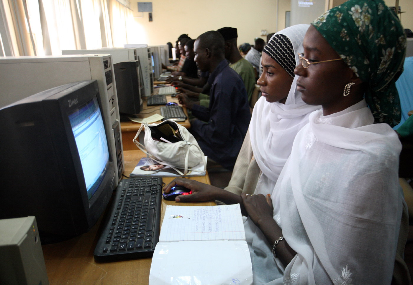 Students working on computers in a university computer lab, including a woman in a green hijab actively using a mouse.