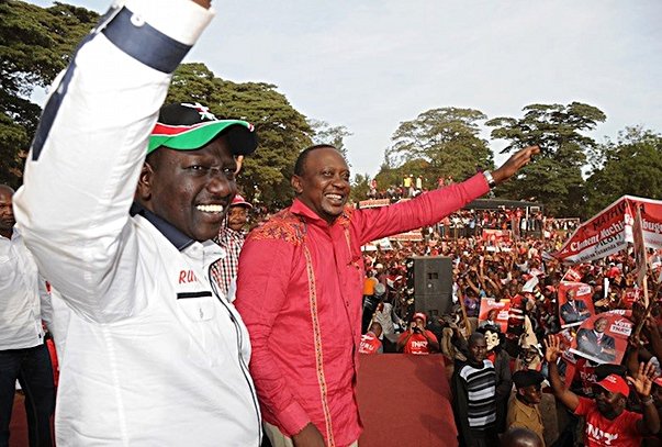 Two politicians in red attire waving to a crowd of supporters at an outdoor rally, with banners and flags visible in the background.