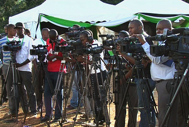 A group of cameramen operating video cameras, standing outdoors with a tent in the background.