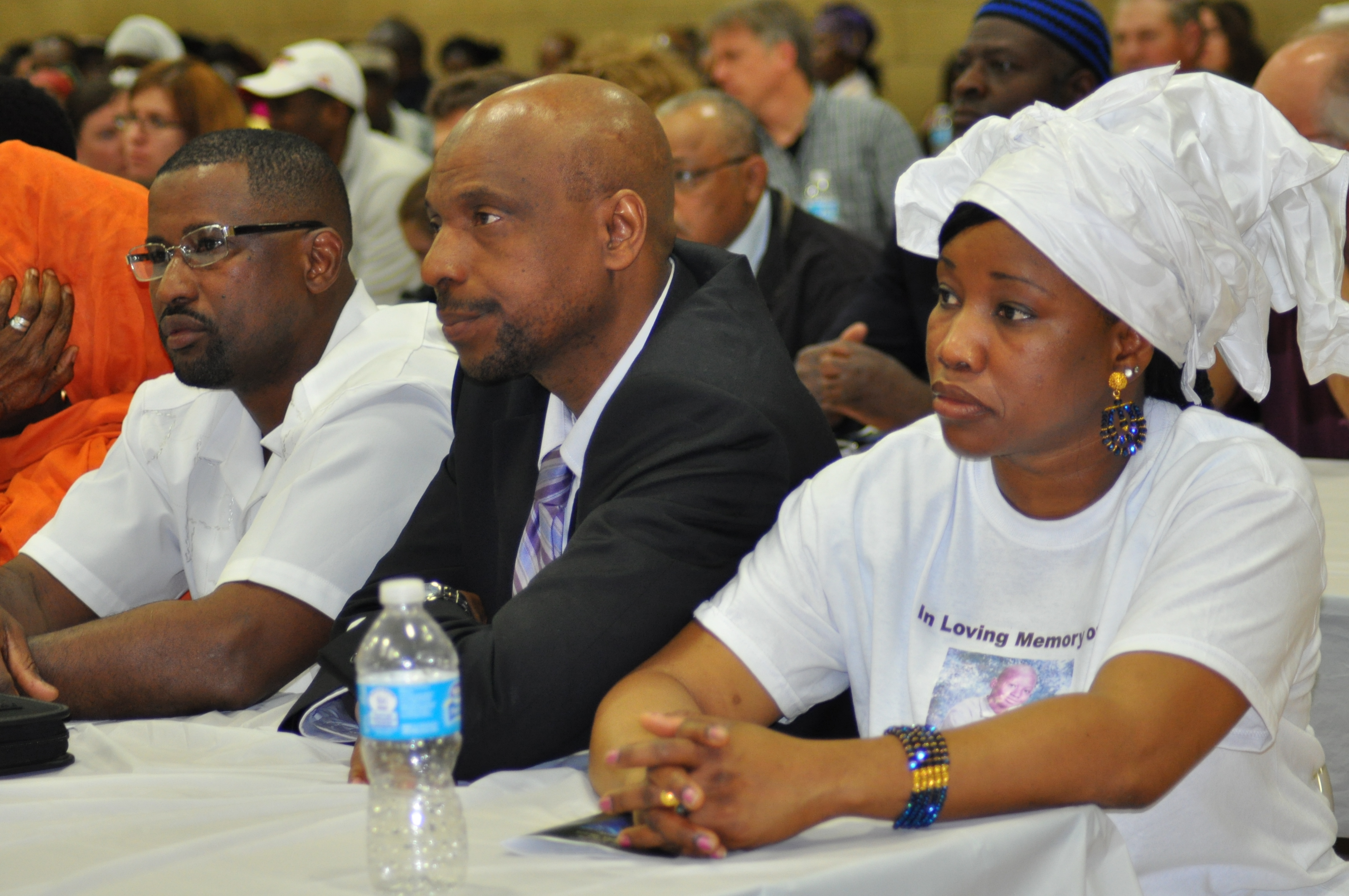 Three adults sitting at a table attentively watching an event, with a woman in a white headwrap on the right.