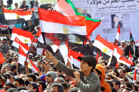 A large crowd of demonstrators waving egyptian flags during a protest, with a young man prominently holding up a flag in the foreground.