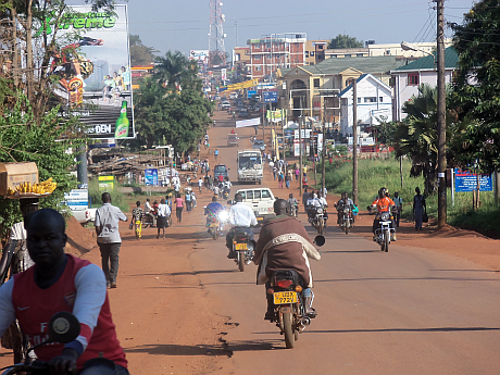 Busy street scene in an african town with motorcycles and pedestrians, surrounded by commercial signs and lush greenery.
