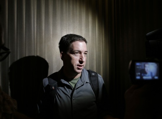 A man in a dark shirt speaking during an interview, lit by a spotlight, with a camera visible in the foreground.