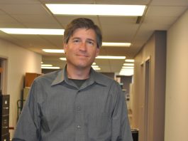 A man in grey shirt standing next to a wall.
