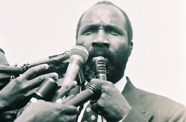 A man in suit and tie holding up several microphones.